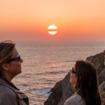Deux femmes et un homme portant des lunettes de protection observent une éclipse solaire partielle au-dessus d’un horizon marin dégagé, au coucher du Soleil, sur une côte française.
