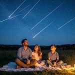Une famille assise sur des couvertures dans une prairie sombre observe émerveillée une pluie d’étoiles filantes sous un ciel nocturne dégagé.